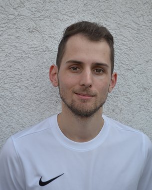 Ein junger Mann mit kurzen Haaren trägt ein weißes T-Shirt mit einem schwarzen Nike-Logo. Er steht vor einer strukturierten, hellen Wand und blickt direkt in die Kamera.