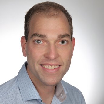 Ein Mann mit kurzen, braunen Haaren und einem freundlichen Lächeln trägt ein hellblaues Hemd mit einem weißen Kragen.