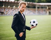 Ein Mann in einem schwarzen Anzug hält einen Fußball in der Hand, während er auf einem Sportfeld steht.