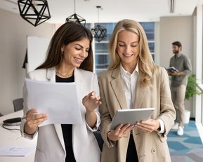 Zwei Geschäftspersonen in Anzügen betrachten gemeinsam ein Tablet in einem modernen Büro. Ein Tisch mit Laptops und Stühlen ist im Hintergrund sichtbar.