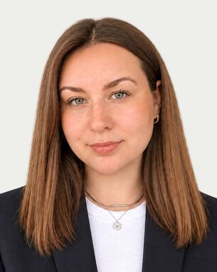 Eine Person mit langen, braunen Haaren und grünen Augen trägt einen schwarzen Blazer und ein weißes Oberteil.