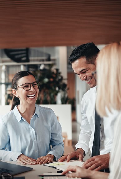 Drei Personen in einem modernen Büro diskutieren und lächeln. Eine Frau mit Brille trägt ein hellblaues Hemd, während die anderen beiden in formeller Kleidung sind. Ein Laptop und Dokumente liegen auf dem Tisch.