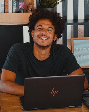 Ein junger Mann mit lockigem Haar sitzt an einem Tisch vor einem Laptop. Im Hintergrund sind Bücherregale und Pflanzen sichtbar. Ein Schild mit dem Text "WIFI PASSWORD" steht ebenfalls auf dem Tisch.