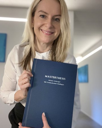 Eine Frau hält stolz ihre Masterarbeit in der Hand. Der Titel "MASTERTHESIS" und ihr Name "Melanie Grunigel" sind auf dem Cover zu sehen. Der Hintergrund ist hell und modern gestaltet.