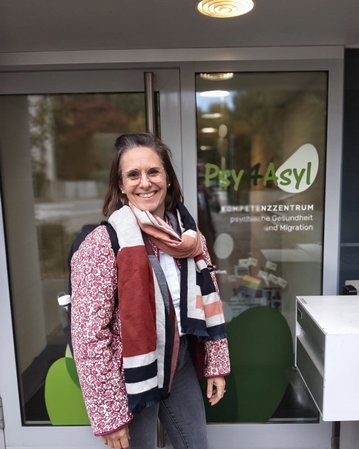 Frau mit Brille und Schal steht vor dem Eingang des Psychiatrie- und Migrationszentrums "Psy 4 Asyl".