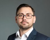 Ein Mann mit Brille trägt einen Anzug und schaut direkt in die Kamera. Sein Gesichtsausdruck ist neutral, und er hat kurzes, dunkles Haar. Der Hintergrund ist einfarbig und unauffällig.