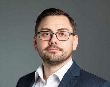 Ein Mann mit Brille trägt einen Anzug und schaut direkt in die Kamera. Sein Gesichtsausdruck ist neutral, und er hat kurzes, dunkles Haar. Der Hintergrund ist einfarbig und unauffällig.