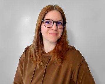 Eine Person mit langen, braunen Haaren und Brille lächelt in die Kamera, trägt einen braunen Hoodie und hat einen neutralen Hintergrund.