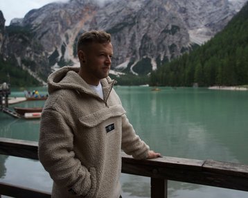 Ein Mann in einem beigen Fleece-Pullover steht an einem Holzgeländer und blickt auf einen grünen See, umgeben von Bergen und Wäldern.
