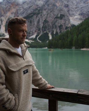 Ein Mann in einem beigen Fleece-Pullover steht an einem Holzgeländer und blickt auf einen grünen See, umgeben von Bergen und Wäldern.