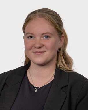 Junge Frau mit blonden Haaren, die in einem schwarzen Blazer lächelt. Der Hintergrund ist neutral und hell.