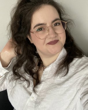 Eine Person mit lockigem, dunklem Haar und Brille lächelt in die Kamera, während sie eine Hand durch ihr Haar fährt. Sie trägt ein weißes Hemd und hat einen neutralen Hintergrund.