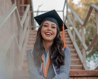 Eine junge Frau mit einem Abschlusshut sitzt auf einer Treppe und lächelt. Sie trägt eine akademische Robe und mehrere Medaillen um den Hals.