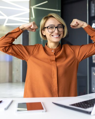 Eine lächelnde Frau mit Brille in einem modernen Büro hebt die Arme in einer Siegerpose. Vor ihr steht ein Laptop auf dem Tisch, daneben liegen ein Notizbuch und Stifte.