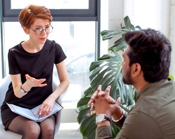 Eine Frau mit Brille sitzt auf einem Stuhl und spricht mit einem Mann, der ihr gegenüber sitzt. Beide scheinen in ein Gespräch vertieft zu sein, umgeben von Pflanzen und natürlichem Licht.