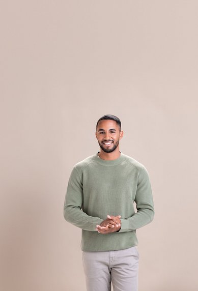 Ein Mann steht lächelnd mit verschränkten Händen in einem hellen, einfarbigen Raum. Er trägt einen grünen Pullover und graue Hosen.