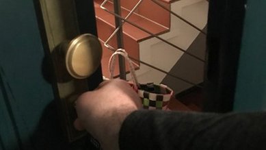Eine Hand greift nach einem Türknauf, während eine Treppe im Hintergrund sichtbar ist. Ein kariertes Einkaufstasche hängt an der Treppe.