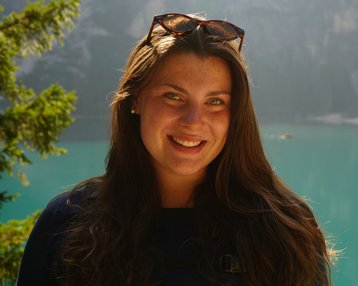 Eine Frau mit langen, braunen Haaren und Sonnenbrille lächelt vor einem klaren, türkisfarbenen See und bewaldeten Bergen.