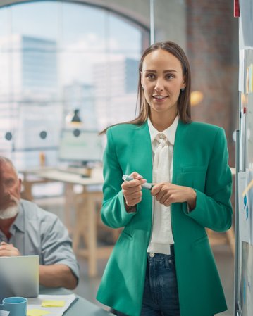 Eine Frau in einem grünen Blazer steht vor einer Gruppe, hält einen Stift und spricht. Im Hintergrund sind Notizen und ein Büro sichtbar.