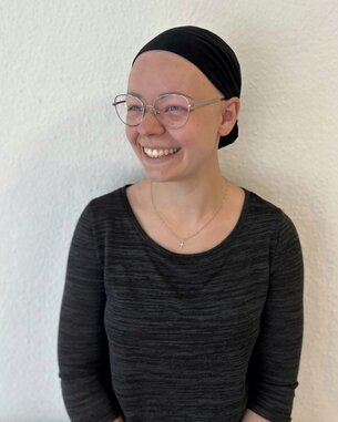 Eine lächelnde Person mit einer schwarzen Kopfbedeckung und Brille, die ein schwarzes Oberteil trägt, steht vor einer weißen Wand.