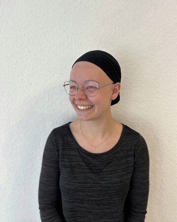 Eine lächelnde Person mit einer schwarzen Kopfbedeckung und Brille, die ein schwarzes Oberteil trägt, steht vor einer weißen Wand.