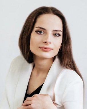 Eine Frau mit langen, braunen Haaren und einem neutralen Ausdruck trägt einen weißen Blazer und posiert vor einem hellen Hintergrund.