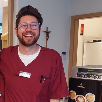 Ein lächelnder Mann in rotem Arztkittel steht neben einer Kaffeemaschine. Er trägt eine Armbanduhr und hat eine Brille auf. Im Hintergrund sind Möbel und eine Wand sichtbar.