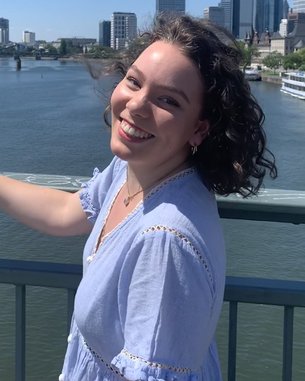 Eine Person mit lockigem Haar steht auf einer Brücke und lächelt, während sie auf einen Fluss und die Skyline einer Stadt im Hintergrund blickt. Es ist ein sonniger Tag.
