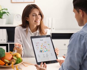 Eine Frau zeigt einem Mann auf einem Tablet eine Ernährungstabelle, während sie am Tisch mit Obst und Gemüse sitzt.
