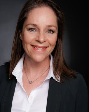 Porträt einer Frau mit langen, braunen Haaren, die ein weißes Hemd und einen schwarzen Blazer trägt. Sie lächelt freundlich in die Kamera.