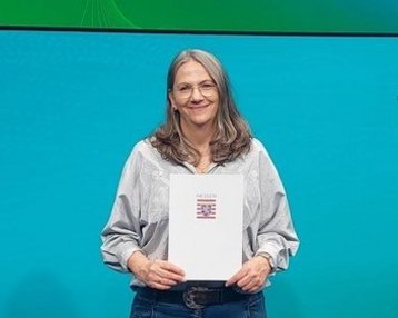 Eine Frau hält eine Urkunde in der Hand und steht vor einem Bildschirm mit dem Text "formnext". Sie trägt ein graues Oberteil und Jeans.