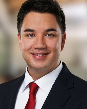 Ein junger Mann mit dunklen Haaren trägt einen Anzug und eine rote Krawatte. Er lächelt und steht vor einem unscharfen, modernen Hintergrund.