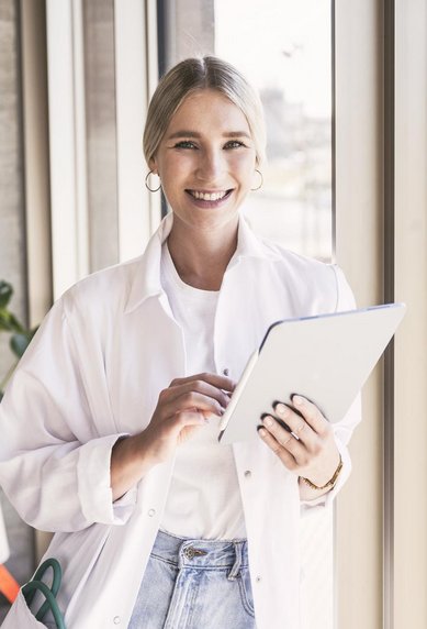 Eine lächelnde Frau in einem weißen Laborkittel hält ein Tablet in der Hand, während sie an einem Fenster steht. Im Hintergrund ist eine Pflanze und ein urbaner Ausblick zu sehen.