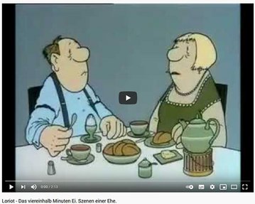 Ein Mann und eine Frau sitzen an einem Tisch, während sie ein Frühstück mit einem Ei, Brötchen und Tee genießen. Der Mann schaut die Frau an, die ernst wirkt.