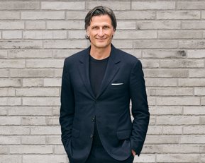 Mann in einem dunklen Anzug steht mit verschränkten Händen vor einer grauen Ziegelwand.