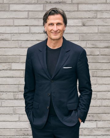 Mann in einem dunklen Anzug steht mit verschränkten Händen vor einer grauen Ziegelwand.