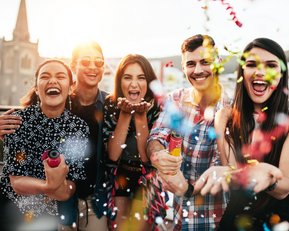 Eine Gruppe von fünf fröhlichen Menschen steht zusammen und wirft Konfetti in die Luft. Sie lächeln und halten Partyartikel in den Händen, während die Sonne im Hintergrund scheint.