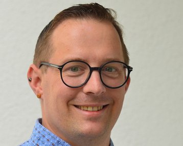 Ein lächelnder Mann mit Brille trägt ein gemustertes Hemd. Er hat kurze, hellbraune Haare und Ohrstecker. Der Hintergrund ist neutral und hell.