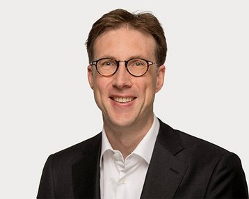 Ein Mann mit Brille und kurzem, braunem Haar trägt einen schwarzen Anzug und ein weißes Hemd. Er lächelt freundlich in die Kamera. Der Hintergrund ist neutral und hell.