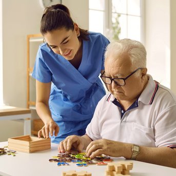 Eine Pflegekraft hilft einem älteren Mann beim Puzzeln am Tisch, während er konzentriert an den Teilen arbeitet.