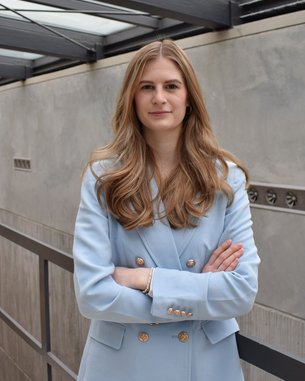 Eine Frau mit langen, lockigen Haaren steht mit verschränkten Armen in einem modernen Innenraum. Sie trägt einen hellblauen Blazer und blickt direkt in die Kamera. Der Hintergrund zeigt eine minimalistische Architektur.