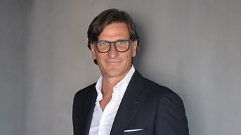 Ein Mann in einem dunklen Anzug mit Brille steht vor einer grauen Wand. Er trägt ein weißes Hemd und hat eine entspannte Haltung.