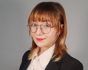 Eine Person mit schulterlangen, braunen Haaren trägt einen schwarzen Anzug und eine weiße Bluse. Sie steht mit verschränkten Händen und schaut direkt in die Kamera.