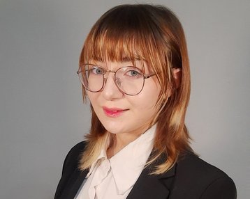 Eine Person mit schulterlangen, braunen Haaren trägt einen schwarzen Anzug und eine weiße Bluse. Sie steht mit verschränkten Händen und schaut direkt in die Kamera.