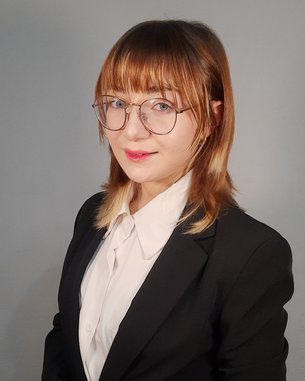 Eine Person mit schulterlangen, braunen Haaren trägt einen schwarzen Anzug und eine weiße Bluse. Sie steht mit verschränkten Händen und schaut direkt in die Kamera.