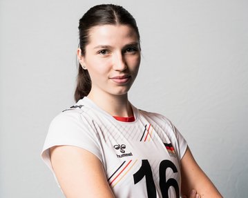 Eine junge Frau in einem weißen Volleyballtrikot mit der Nummer 16 steht mit verschränkten Armen vor einem neutralen Hintergrund. Sie hat lange, braune Haare und schaut direkt in die Kamera.