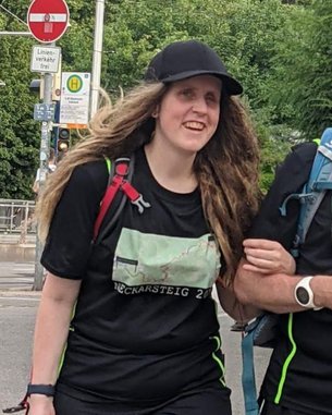 Ein Mann und eine Frau in sportlicher Kleidung mit Rucksäcken gehen zusammen auf einem Gehweg. Beide tragen T-Shirts mit dem Aufdruck "Neckarsteig 2022".