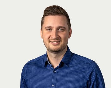 Ein Mann mit kurzen Haaren trägt ein blaues Hemd mit Punkten und lächelt freundlich in die Kamera.