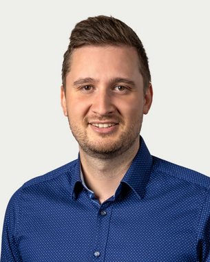 Ein Mann mit kurzen Haaren trägt ein blaues Hemd mit Punkten und lächelt freundlich in die Kamera.