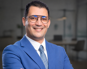 Ein Mann in einem blauen Anzug mit Brille lächelt, während er mit verschränkten Armen vor einem modernen Bürohintergrund steht.